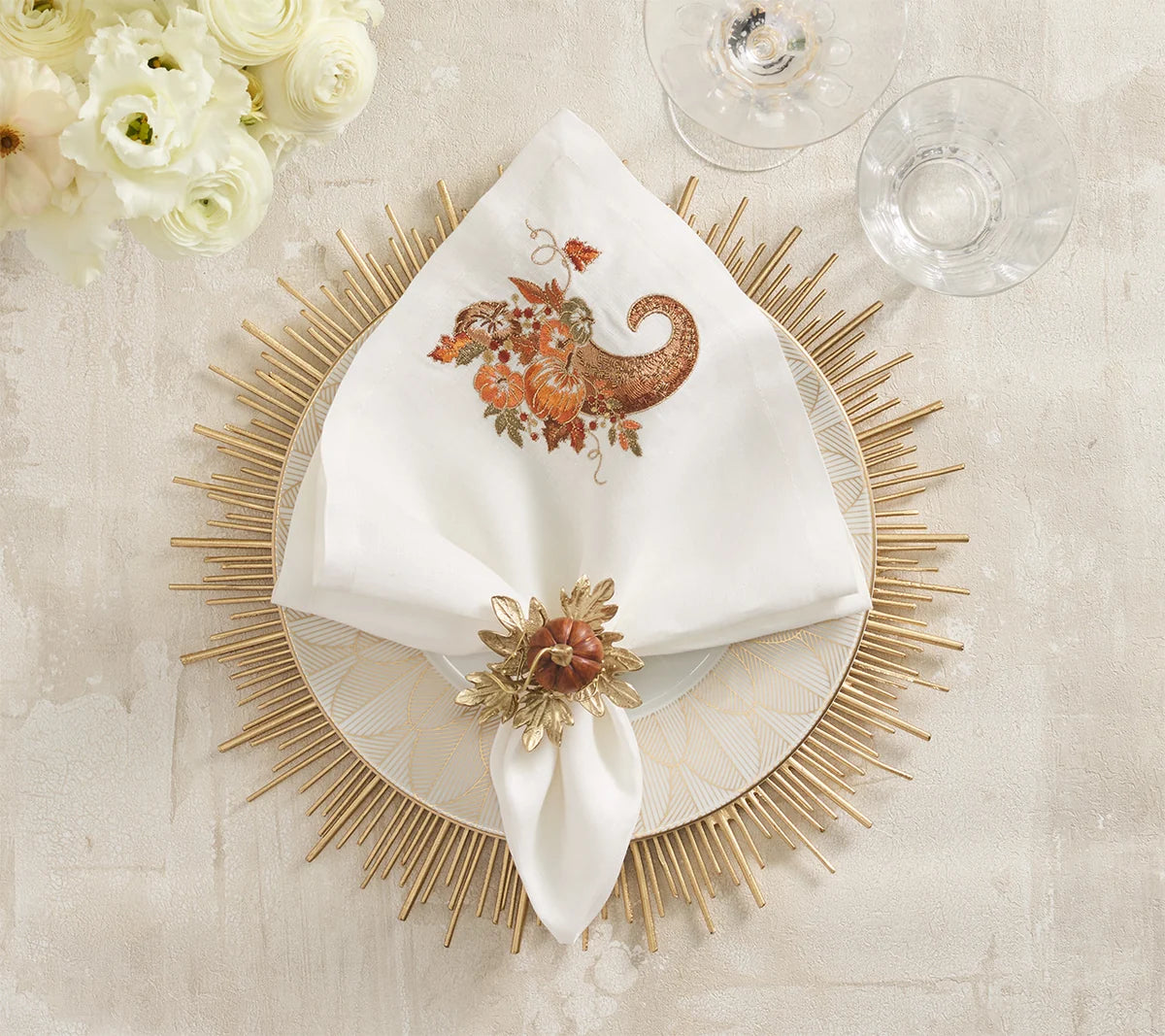 White dinner napkin with an embroidered cornucopia and pumpkins, secured by a Kim Seybert Gilded Gourd Napkin Ring on a decorative charger plate for an elegant fall tablescape.