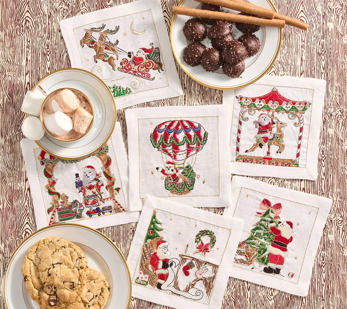 Six Kim Seybert Santa Cocktail Napkins with embroidered festive scenes of Santa Claus, arranged on a wood-grain table with plates of cookies and a mug of hot cocoa.