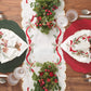 A Kim Seybert Tinsel Bow Table Runner with embroidered red ribbons and green foliage, centered between two place settings with green and red chargers, holiday napkins, and glassware on a wood table.