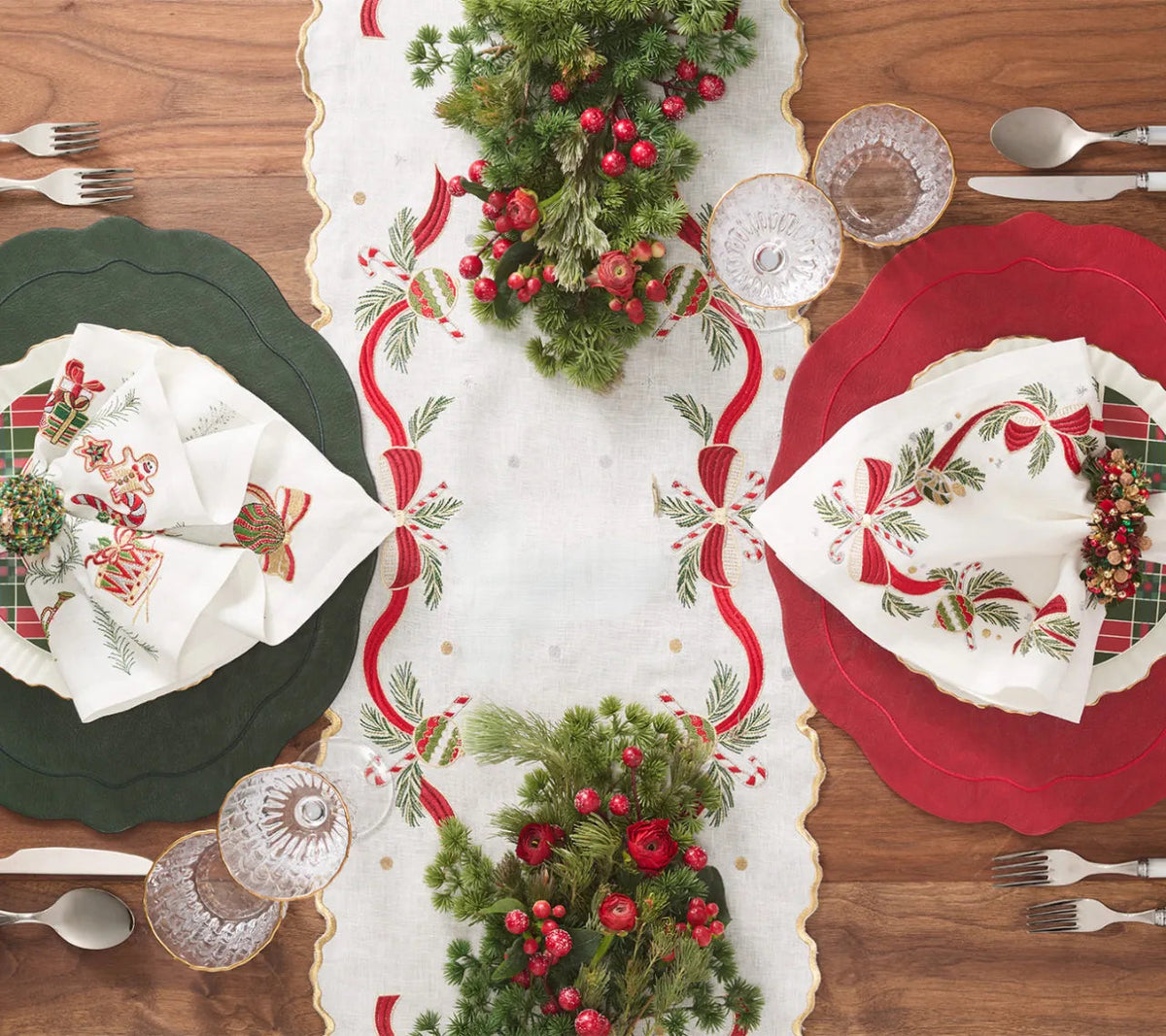 A Kim Seybert Tinsel Bow Table Runner with embroidered red ribbons and green foliage, centered between two place settings with green and red chargers, holiday napkins, and glassware on a wood table.