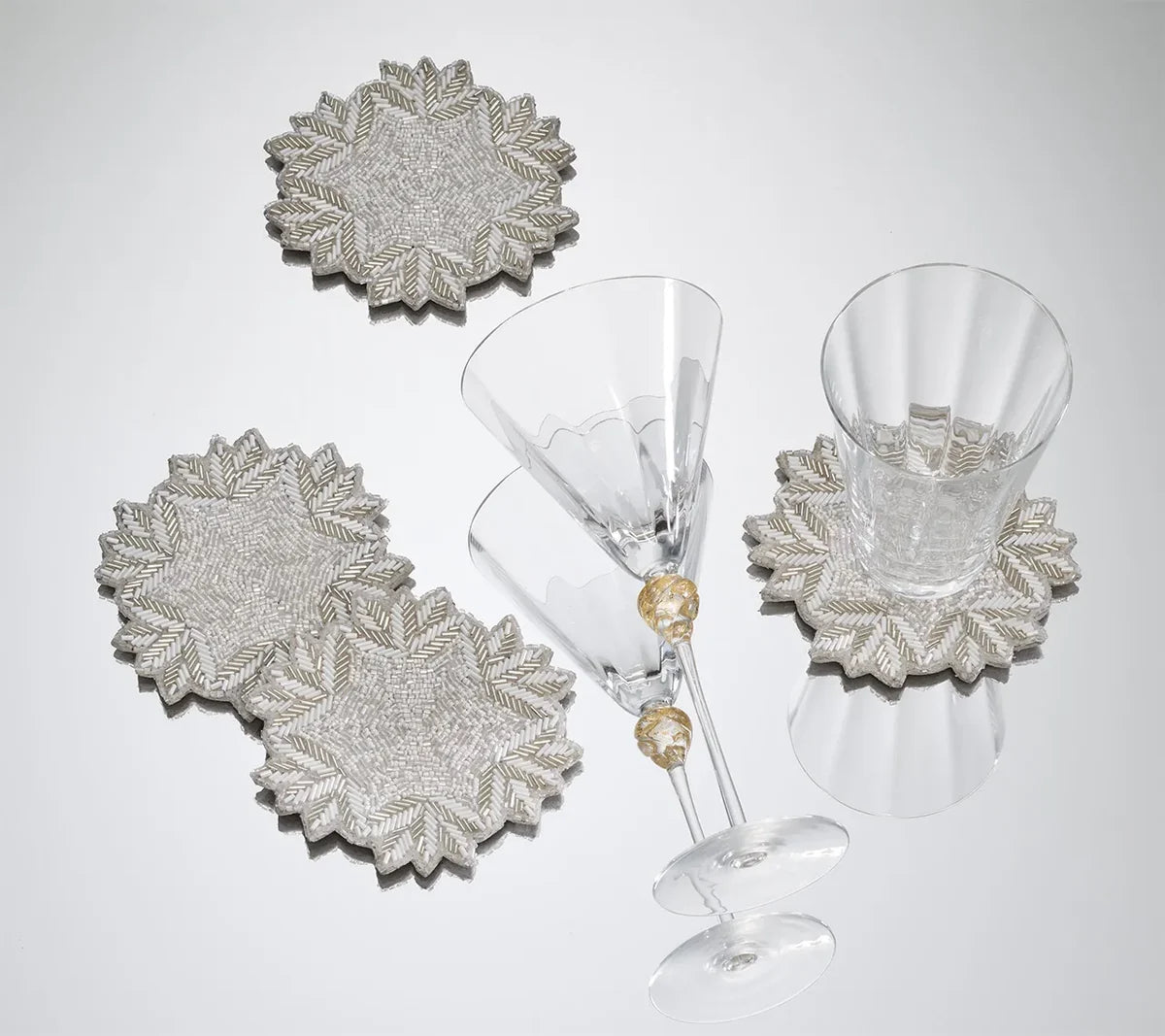 Four silver-beaded Winter Lace Coasters with round, leaf-like edges, two of which are topped with clear glass drinkware.