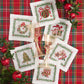 Six Christmas Trellis Cocktail Napkins featuring embroidery of a wreath, tree, ornament, gifts, gingerbread man, and drum, arranged on a red plaid tablecloth with two glass goblets.