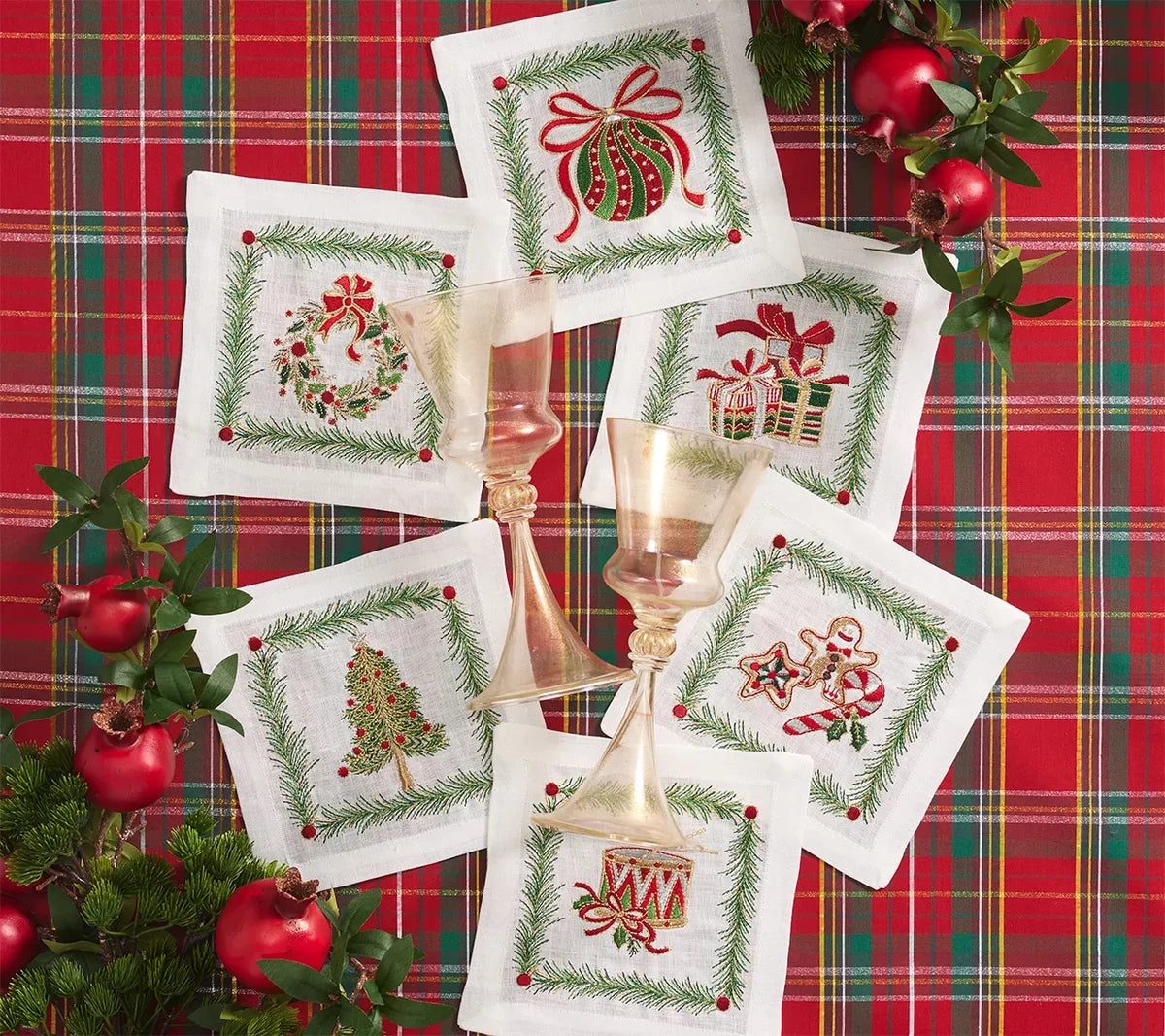 Six Christmas Trellis Cocktail Napkins featuring embroidery of a wreath, tree, ornament, gifts, gingerbread man, and drum, arranged on a red plaid tablecloth with two glass goblets.