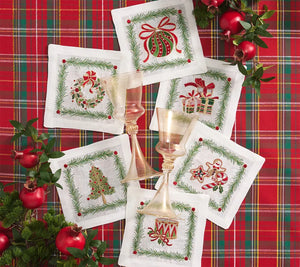 Six Christmas Trellis Cocktail Napkins featuring embroidery of a wreath, tree, ornament, gifts, gingerbread man, and drum, arranged on a red plaid tablecloth with two glass goblets.