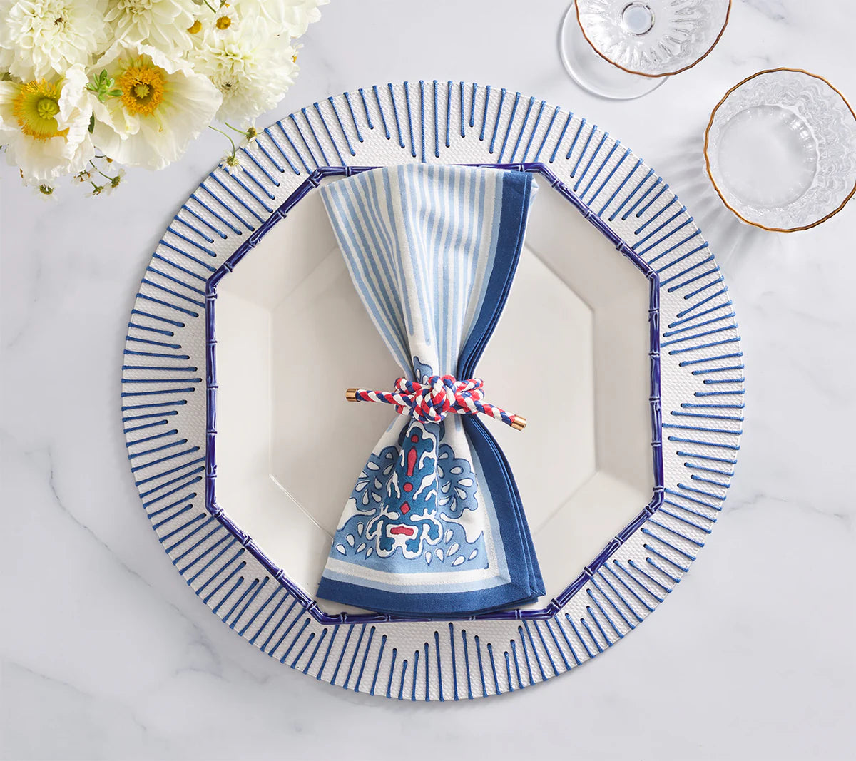 The Kim Seybert Filigree Napkin Set is displayed atop a white octagonal plate with blue trim, accented by a braided cord and surrounded by flowers and glassware for an artisanal touch.