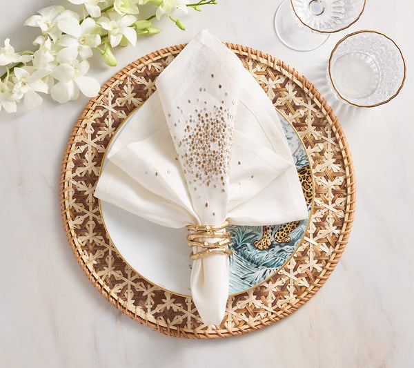 A white napkin with gold accents in a gold napkin ring rests on a decorative plate atop the Kim Seybert Lanai Placemat Collection, set beside clear glasses and white flowers on a marble surface.