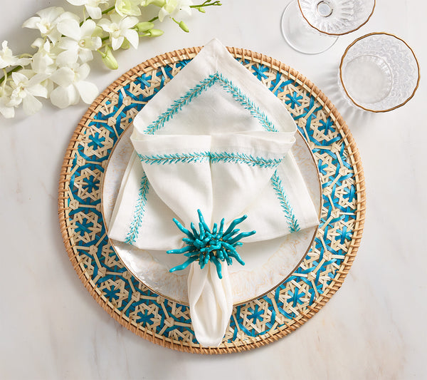 Table setting featuring a Kim Seybert Lanai Placemat, blue and beige patterned plate, white napkin with blue embroidery, blue napkin ring, two glass cups, and white flowers on a marble surface.