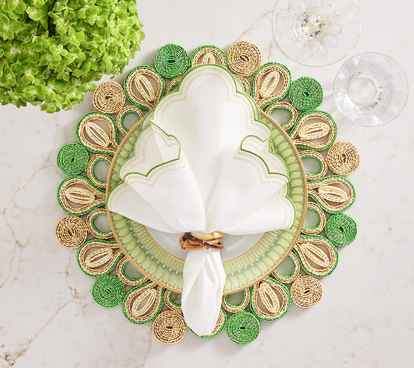 A decorative table setting features a white napkin in a gold ring on a Kim Seybert Marisol Placemat, with a white plate, two clear glasses on marble, and a green floral arrangement.