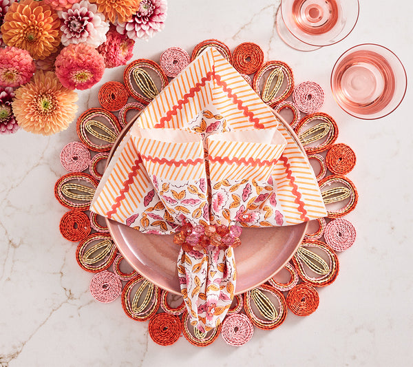 A pink plate with layered napkins, a decorative napkin ring, and a Kim Seybert Marisol Placemat Collection mat, surrounded by flowers and two glasses of pink drink on a marble surface.