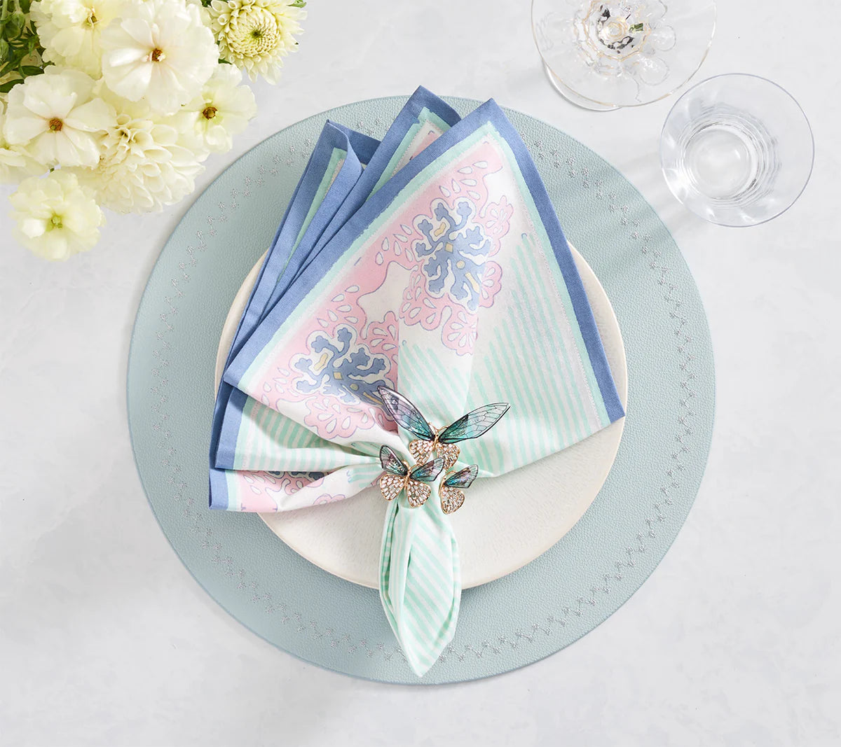 The Kim Seybert Filigree Napkin Set features a folded pastel cotton napkin and decorative filigree ring on a white plate with light blue placemat, glass, and white flowers—showcasing exquisite artisanal craftsmanship.