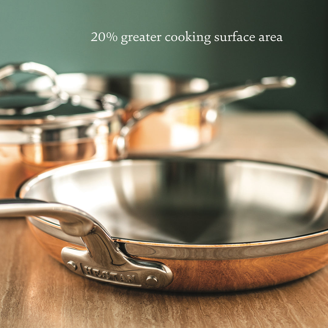 Close-up of the Hestan CopperBond 10-Piece Set’s shiny metal frying pan with 5-ply construction on a wooden surface, plus text above reading "20% greater cooking surface area.