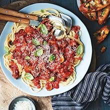 A plate of spaghetti topped with tomato sauce, fresh basil leaves, and grated cheese—served with a fork, spoon, garlic bread, and extra cheese on the side—is just one of the delicious meals you’ll find in Super Easy Pasta! from Simon & Schuster, the perfect choice for anyone seeking quick pasta dishes or easy recipes to enjoy any night of the week.