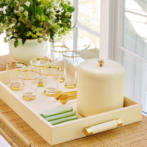 The Aerin Cream Shagreen Rectangular Tray elegantly holds four gold-rimmed glasses, a matching ice bucket, green napkins, and gold tongs; behind it, a floral arrangement sits on a wicker table near the window.