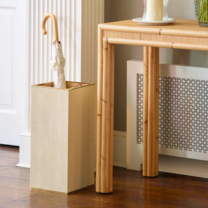 A beige umbrella stands upright in the Aerin Classic Shagreen Umbrella Stand, featuring brass detailing, beside a light woven table on a wooden floor.