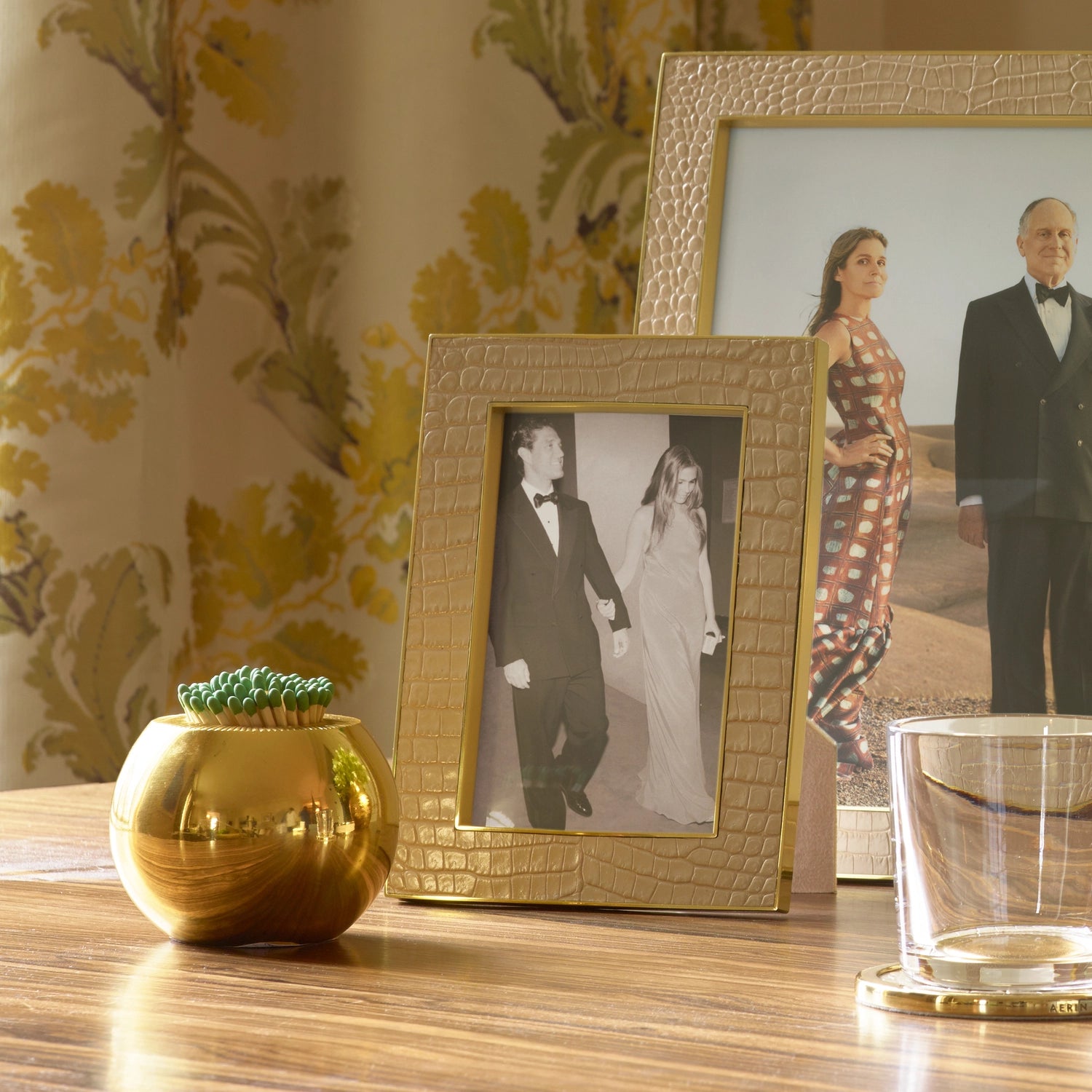 A small gold vase, a glass, an Aerin Sphere Match Striker, and two framed photos sit on a wooden table in front of floral curtains.