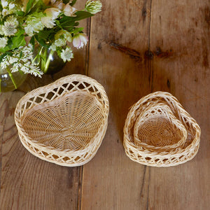 The Aerin Wicker Heart Basket Set features two handcrafted heart-shaped baskets in different sizes, displayed on a rustic wooden table with a vase of white and green flowers adding charm to the scene.