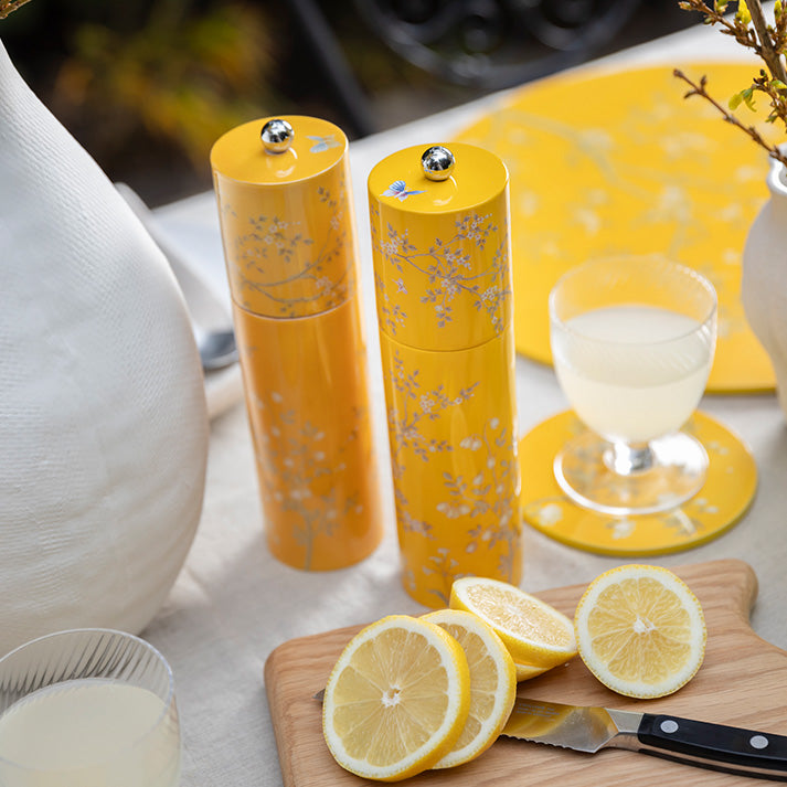 Two yellow floral-patterned mills from the Addison Ross Chinoiserie Salt and Pepper Mill Collection, featuring rubber wood and ceramic grinders, stand on a table beside lemonade, a cutting board with lemons, and a knife.
