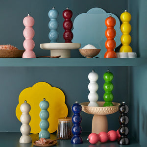 The Hive Bobbin Salt & Pepper Grinder Set is displayed on shelves alongside bowls, small dishes, a jar of peppercorns, and vibrant decorative backgrounds of various shapes and colors.