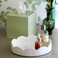 An Addison Ross Lacquer Tissue Box Collection, a green glass vase with foliage, and a white tray with two bottles rest on a beige surface before leafy wallpaper.