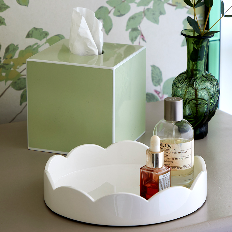 An Addison Ross Lacquer Tissue Box Collection, a green glass vase with foliage, and a white tray with two bottles rest on a beige surface before leafy wallpaper.