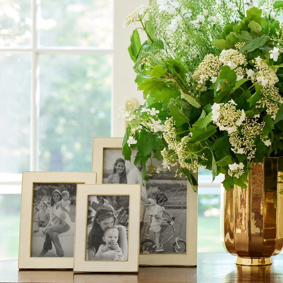 A gold vase with white and green flowers sits beside three Aerin Cream Classic Shagreen photo frames on a table by the window, creating a timeless and elegant display.