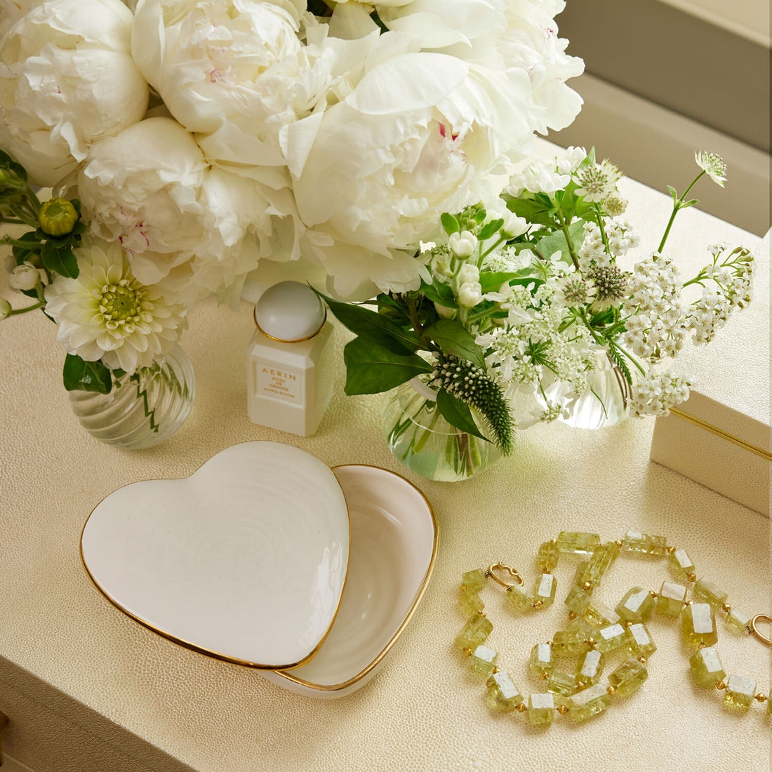 Aerin’s Cream Calinda Heart Box, a floral necklace, two white flower vases, and a perfume bottle are arranged on a cream-colored surface with gold detailing.