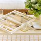 Aerin’s Lacquer Backgammon Set in Cream, with playing pieces and dice, is displayed on a beige table alongside two glasses, a vase of green and white flowers, and a closed notebook.