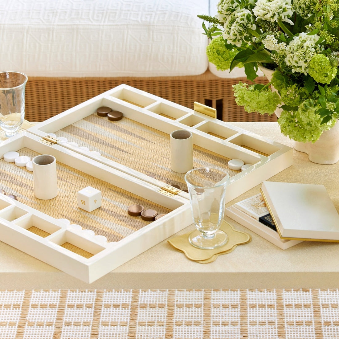 Aerin’s Lacquer Backgammon Set in Cream, with playing pieces and dice, is displayed on a beige table alongside two glasses, a vase of green and white flowers, and a closed notebook.