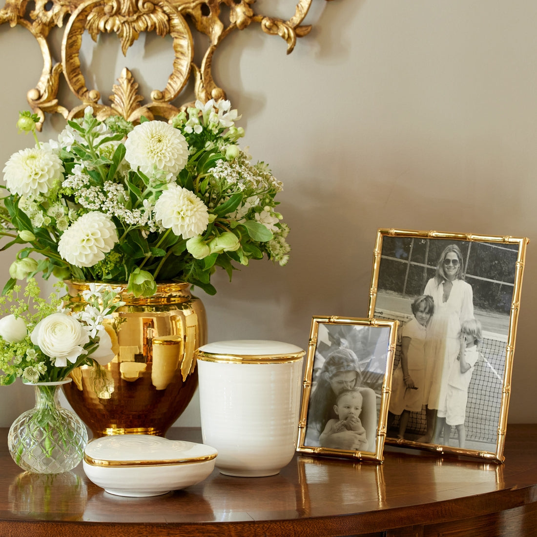 A wooden table displays a gold finish vase with white flowers, two gold-trimmed white containers, a small glass vase, and two black-and-white family photos in Aerin's Ava Bamboo Frame Collection.