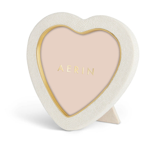 The AERIN Cream Shagreen Heart Frame by Aerin features a heart-shaped design with a gold-bordered, white textured exterior, beige background displaying "AERIN," brass detailing for luxury, and a stand at the back.