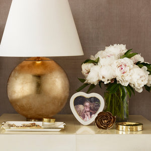 A gold lamp, white peonies in a vase, a gold rose ornament, jewelry on a tray, and Aerin’s Cream Shagreen Heart Frame are displayed on an embossed cream shagreen surface.
