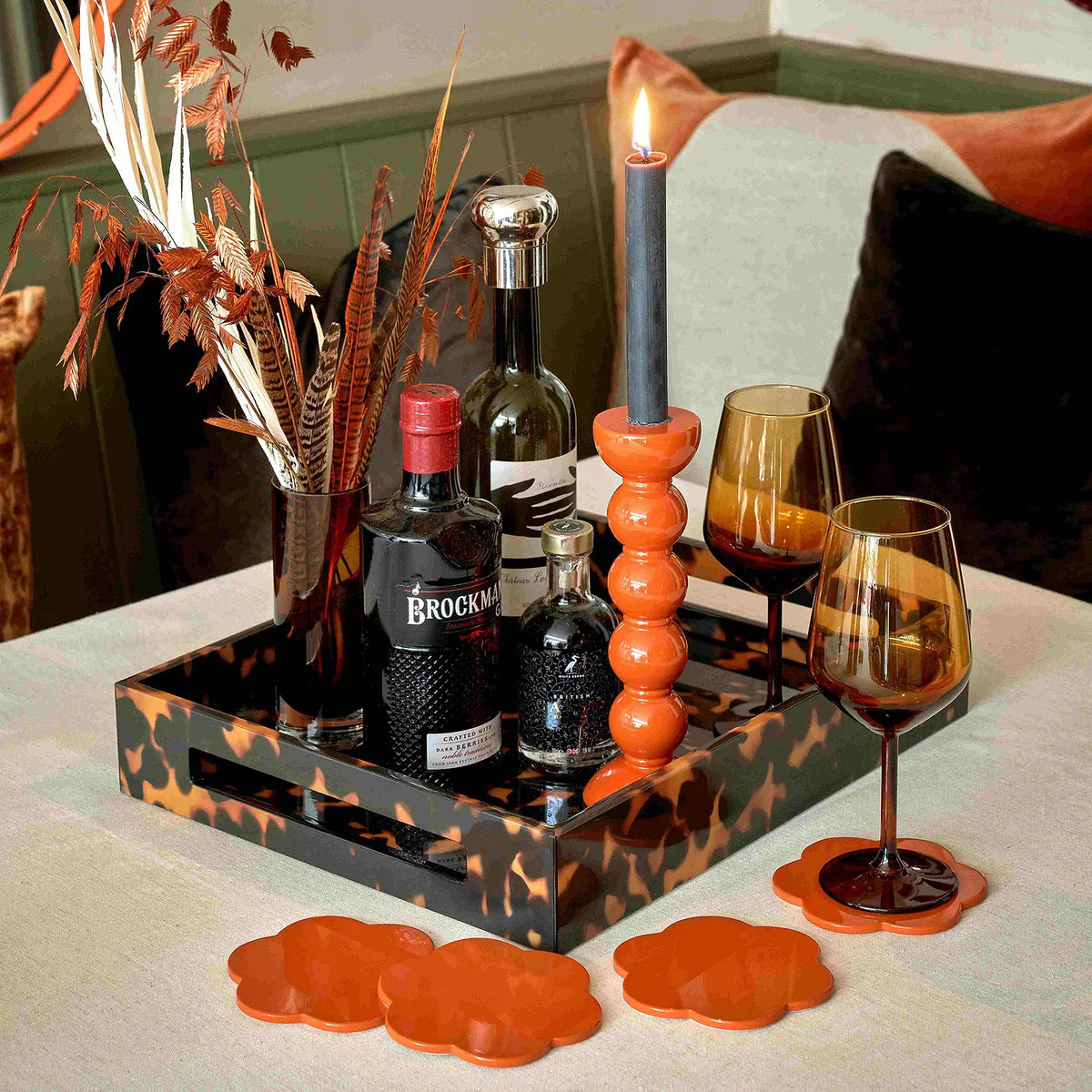 An Addison Ross Faux Tortoise Tray elegantly displays a bottle of Brockmans gin, a glass decanter, a candle, two amber wine glasses, dried leaves in a vase, and orange coasters on a beige tablecloth.