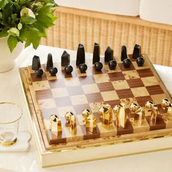 The Aerin Shagreen Chess Set Collection features gold-plated and matte black pieces on a brown and beige board, elegantly showcased on a luxurious shagreen box beside a glass and green plant.