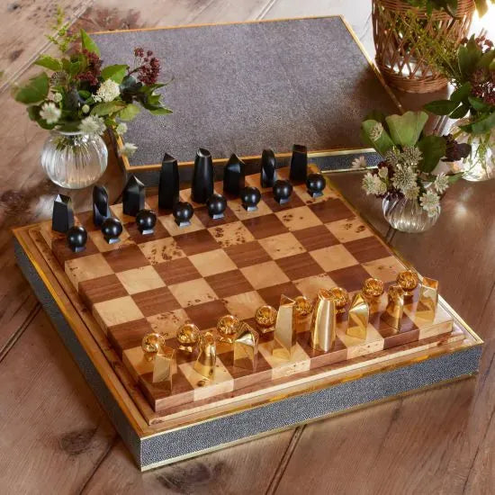 The Aerin Shagreen Chess Set Collection features a wooden chessboard with modern black and gold-plated pieces, complemented by a luxurious shagreen box and small flower vases on the table.