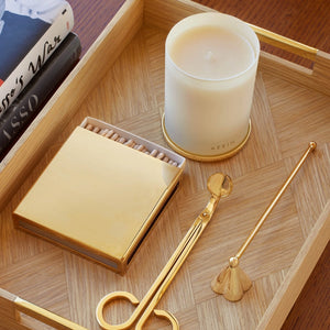 A wooden tray holds a candle, the Aerin Lou Wick Trimmer for candle care, a gold matchbox with matches, a gold candle snuffer, and a book.
