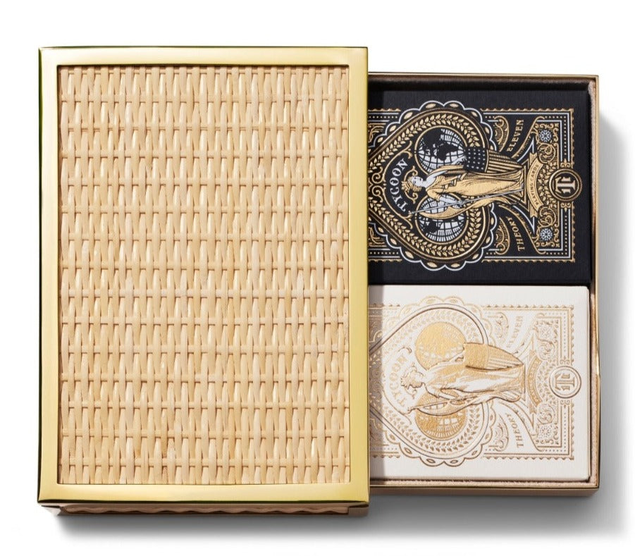 The Aerin Colette Cane Card Set features a rectangular woven box with a gold frame and brass hardware, partially open to display black and white ornate playing card decks resting on a luxurious suede interior.