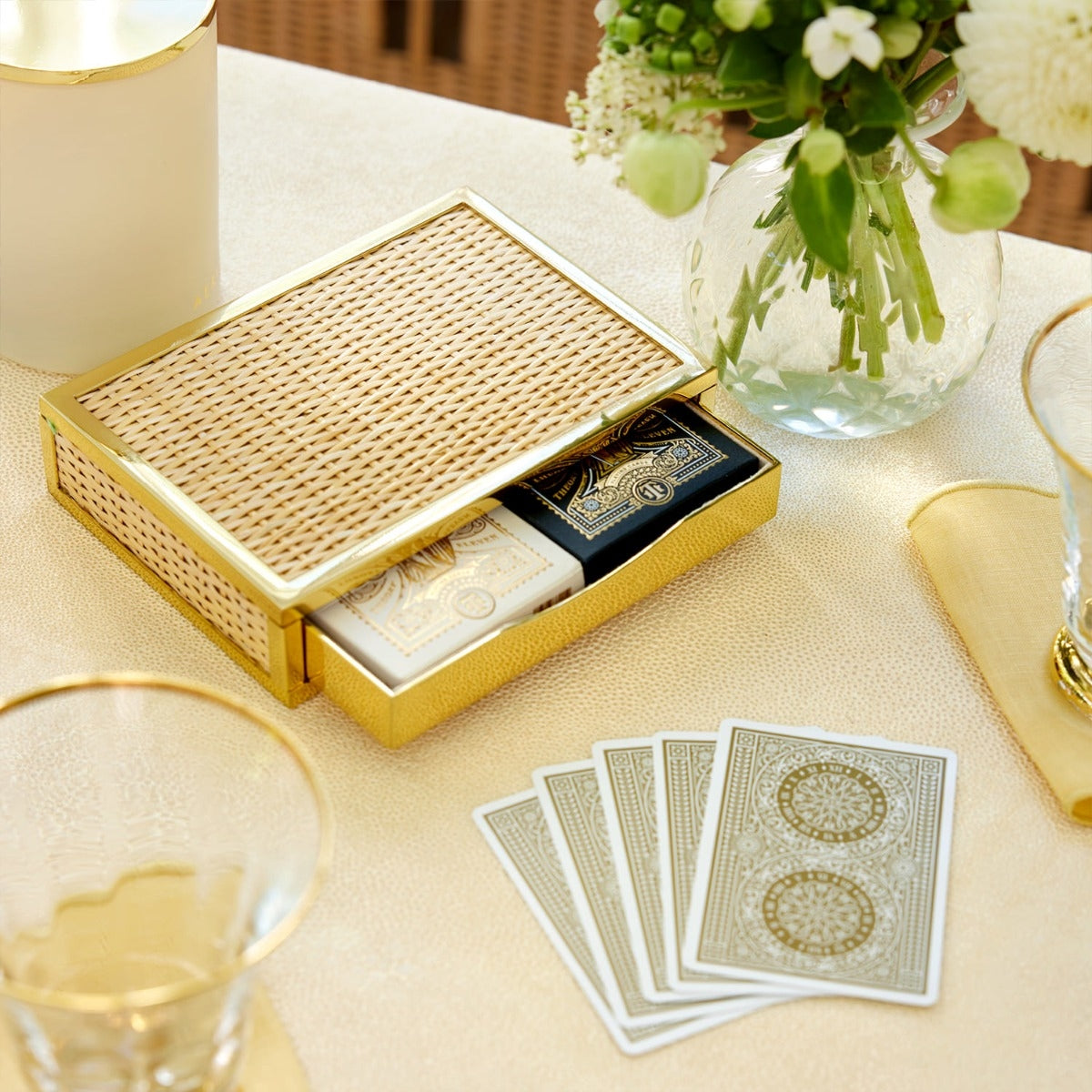 The Aerin Colette Cane Card Set features two decks in a gold and wicker box with a suede interior, displayed on a table beside four cards, glassware, and a vase of white flowers.