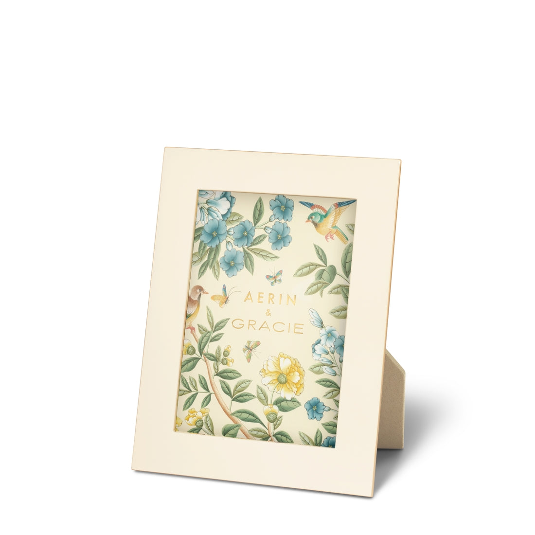 A photo frame from the AERIN x Gracie Heathcote Frame Collection in beige stands on a flat surface, showcasing a floral and bird-illustrated insert with the text "Aerin & Gracie" in the center; it evokes hand-painted wallpapers that elevate traditional interiors.
