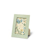 A picture frame from the AERIN x Gracie Heathcote Frame Collection stands on a beige easel backing. The green frame features blue and yellow floral designs that evoke traditional interiors, with the text "Aerin & Gracie" inside.