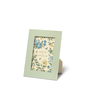 A picture frame from the AERIN x Gracie Heathcote Frame Collection stands on a beige easel backing. The green frame features blue and yellow floral designs that evoke traditional interiors, with the text "Aerin & Gracie" inside.