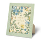 A green picture frame from the AERIN x Gracie Heathcote Frame Collection, displaying floral artwork with birds and butterflies, reminiscent of hand-painted wallpapers.