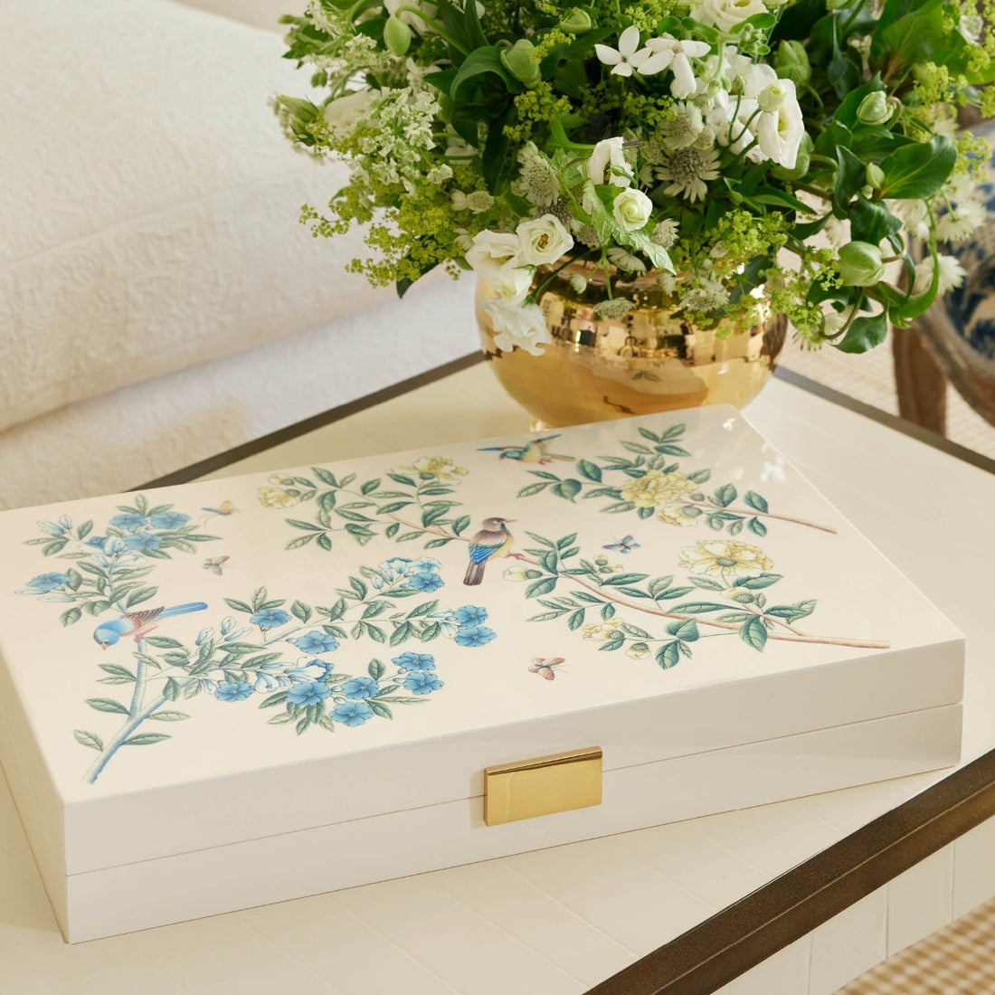 The AERIN x Gracie Heathcote Lacquer Backgammon Set, with floral and bird motifs, complements a gold vase filled with white and green flowers on the table, reflecting Heathcote's elegant design.