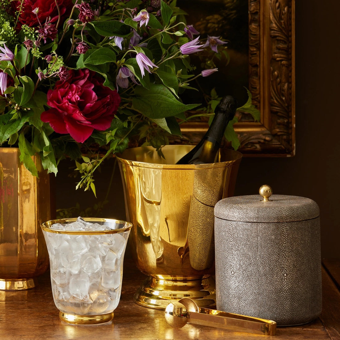 AERIN's Shagreen Ice Bucket in Chocolate is displayed on a wooden surface alongside a bottle, a glass of ice, a textured gray canister with spoon, and a vase of flowers.