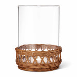 The Navona Rattan Hurricane Collection by Aerin features a clear cylindrical glass vase with a woven rattan pedestal base, adding natural elegance to any space.