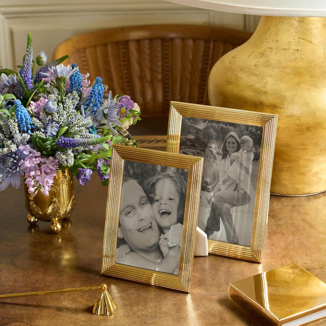 Two photos in gold Alessio frames from Aerin, featuring a reeded design, are displayed with a vase of purple flowers on a wooden table beside a gold lamp and box, creating an elegant arrangement.