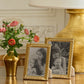 Two Aerin Loretta Frame Collection gold frames with black and white family photos are displayed on a wooden table next to a gold vase with pink flowers and a large gold lamp, creating an elegant photo display.