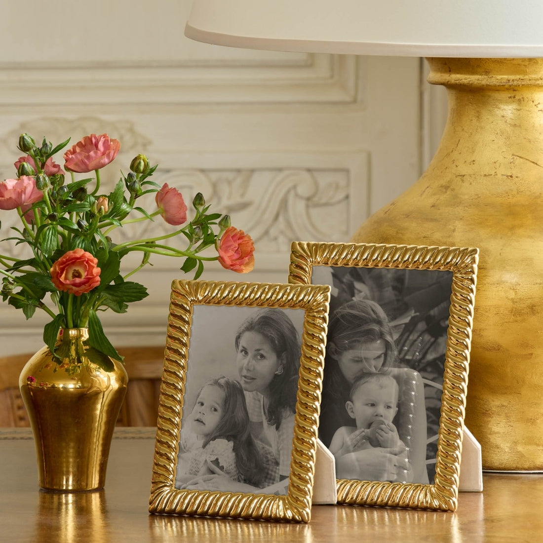 Two Aerin Loretta Frame Collection gold frames with black and white family photos are displayed on a wooden table next to a gold vase with pink flowers and a large gold lamp, creating an elegant photo display.