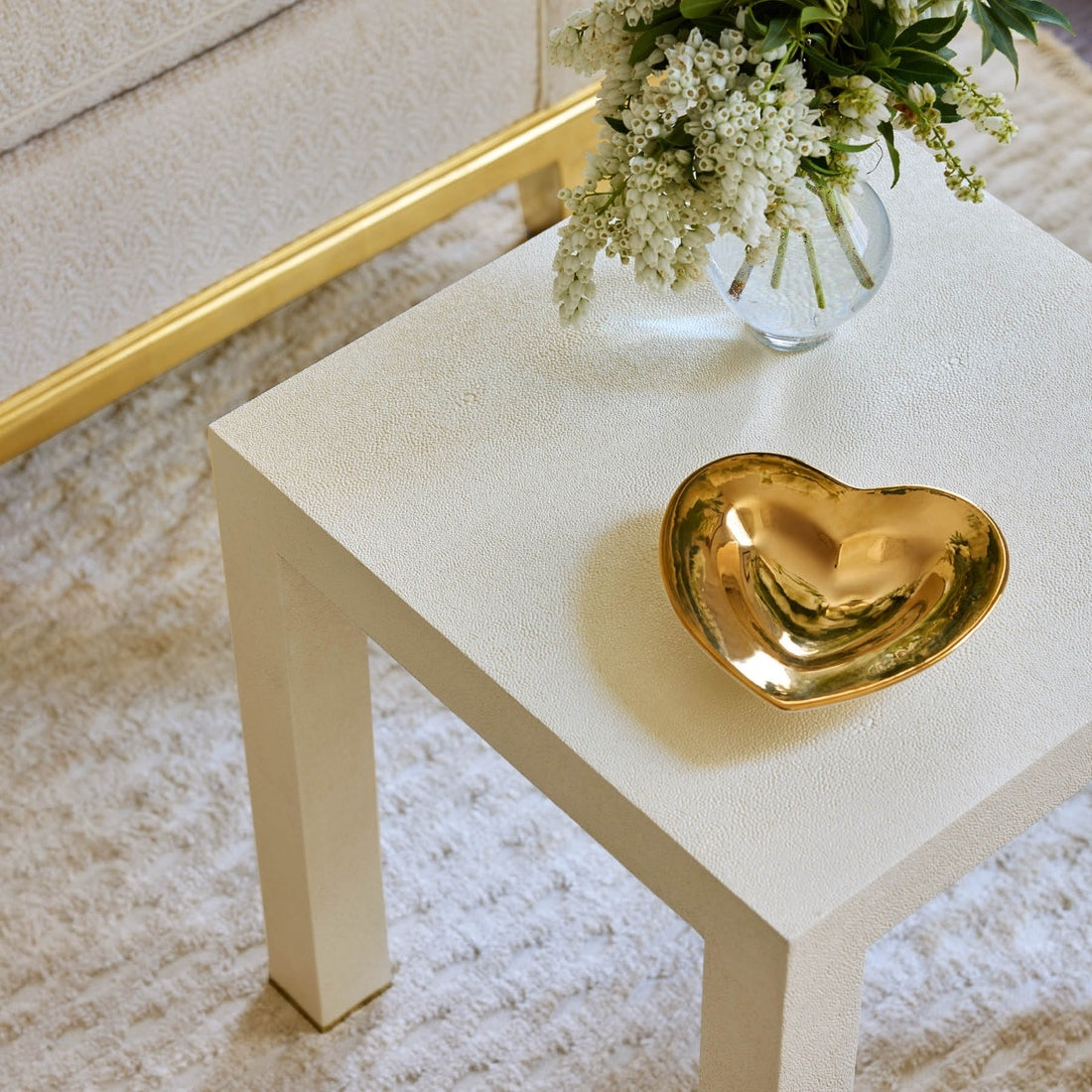 A small white square table features the Aerin Gilded Heart Dish and a glass vase with white flowers, set atop a light textured rug.