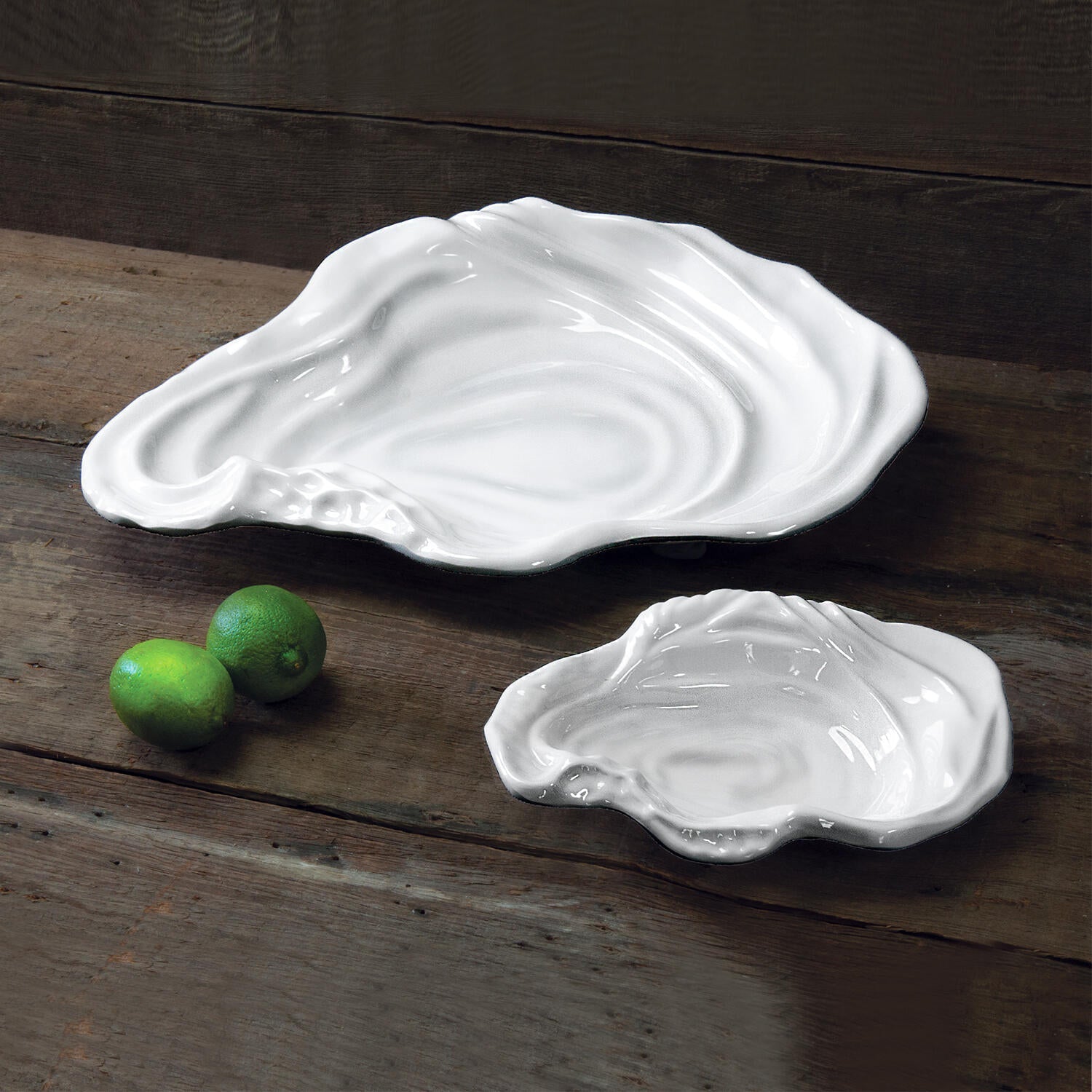 The Beatriz Ball Vida Ocean Oyster Bowl Collection features two white luxury melamine oyster-shaped dishes, placed on a rustic wooden surface with two whole limes—ideal for stylish indoor and outdoor entertaining.