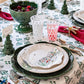 A festive table setting showcases Juliska's Berry & Thread North Pole Dinnerware and Serveware Collection, alongside holiday glassware, red napkins, small tree figurines, and a patterned tablecloth.